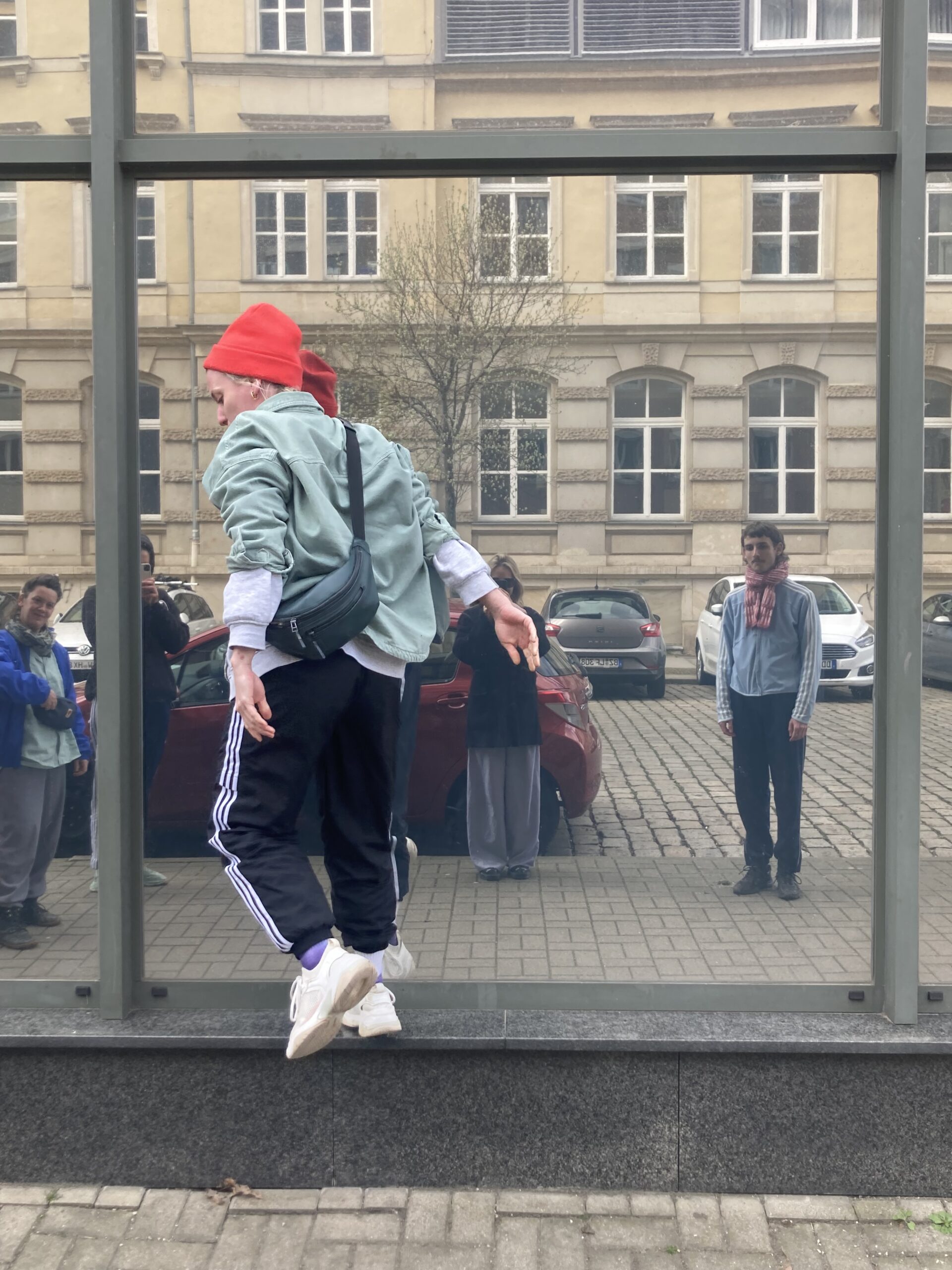 Frau mit roter Mütze vor einer spiegelnden Fassade, Zuschauende in der Spiegelung