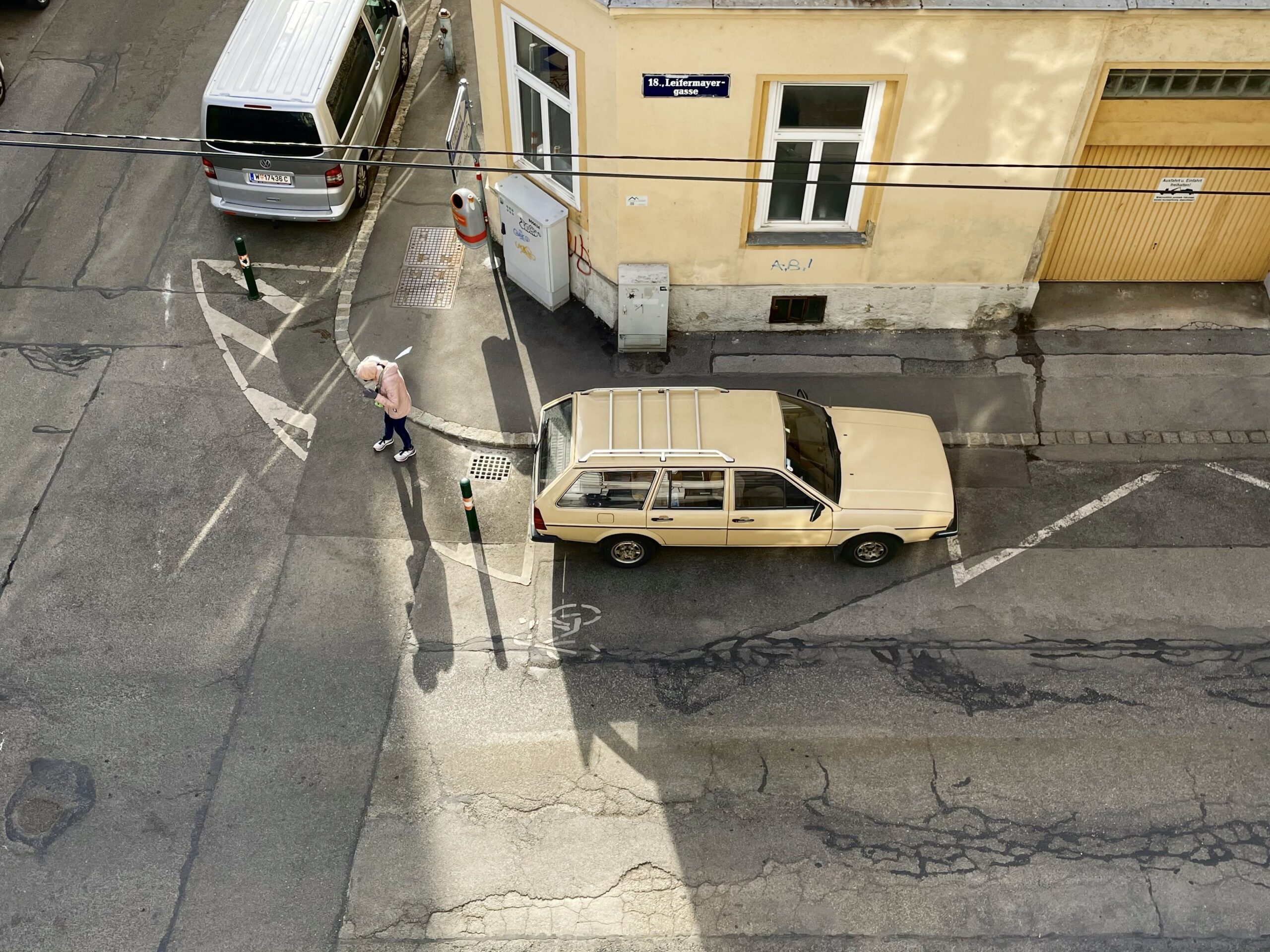 Hellgelber Oldtimer vor hellgelmem Altbau, rosa gekleidete ältere Person geht über Straßenmarkierungen