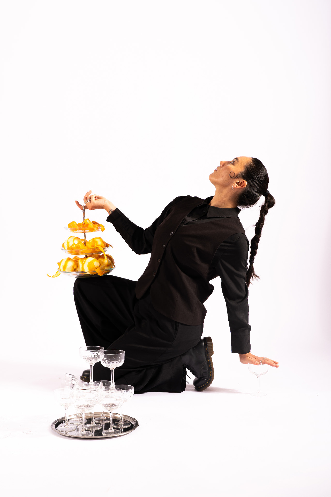 Eine Person kniet auf dem Boden, hält eine Etagere mit Orangenschalen und blickt nach oben, umgeben von Gläsern.